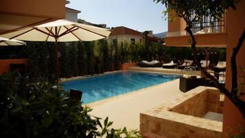 A swimming pool with lounge chairs and umbrellas, surrounded by greenery and buildings.
