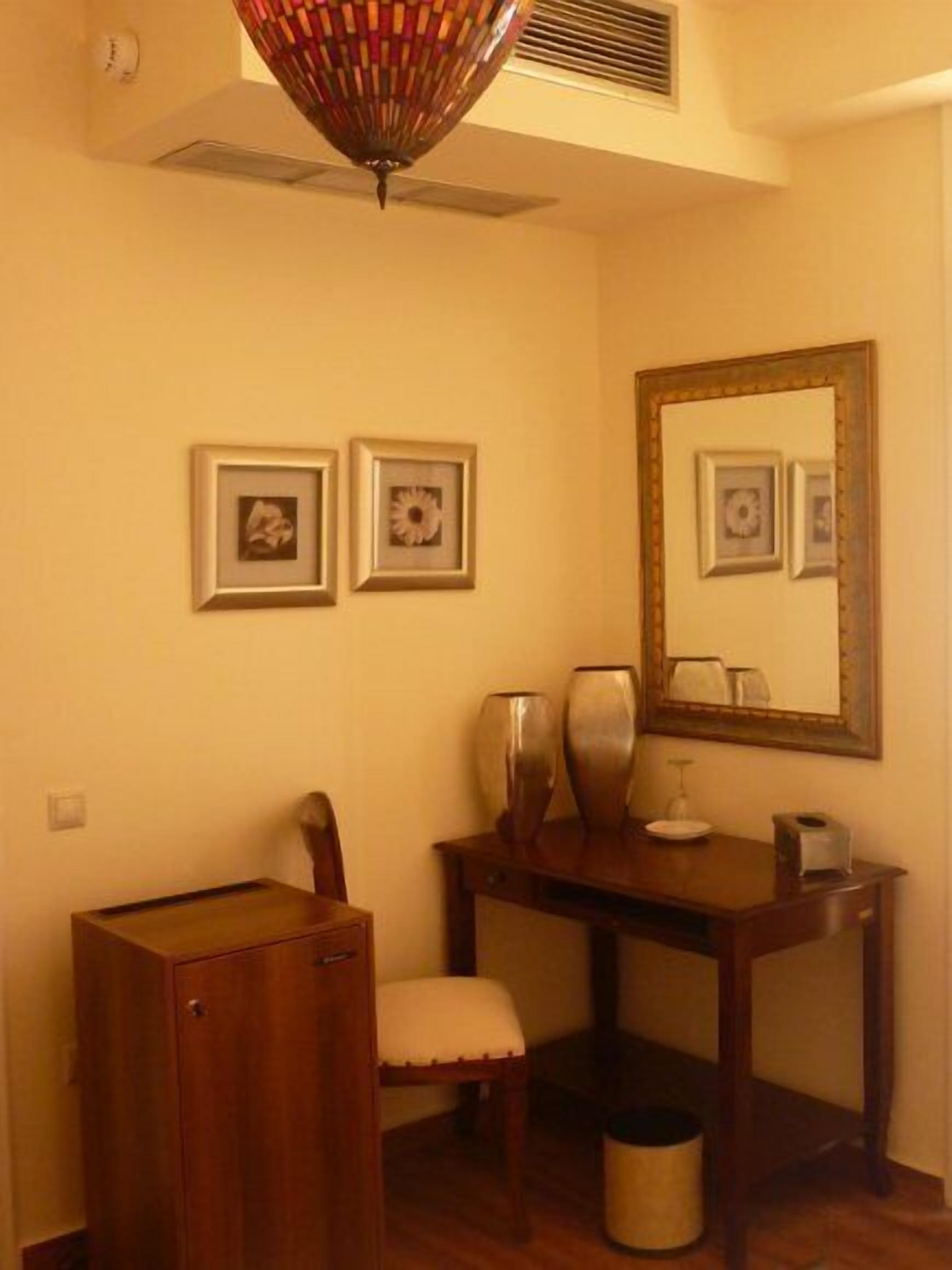 Una habitación con un escritorio de madera, una silla, un mueble pequeño y un cubo de basura. Hay tres cuadros enmarcados en la pared y un espejo sobre el escritorio.