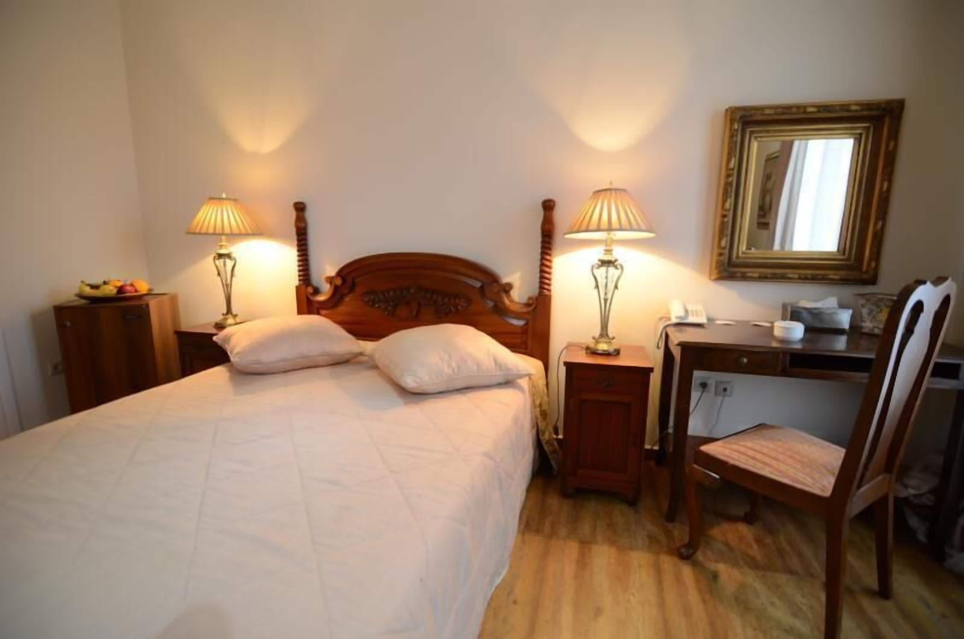 A bedroom with a wooden bed, two bedside lamps, a desk with a chair, and a mirror on the wall.
