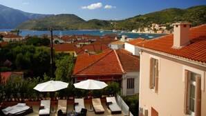 A coastal town with red-roofed buildings, a view of the sea, and a patio with lounge chairs and umbrellas. - Korina Gallery Hotel (Ithaki)