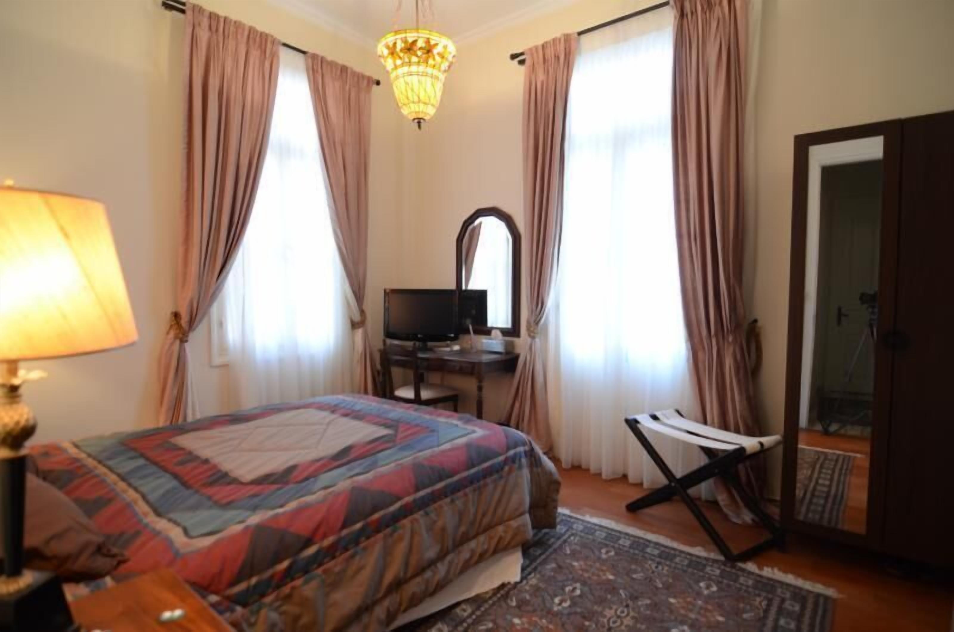 A bedroom with a bed, a desk with a computer, a chair, a mirror, and a chandelier.