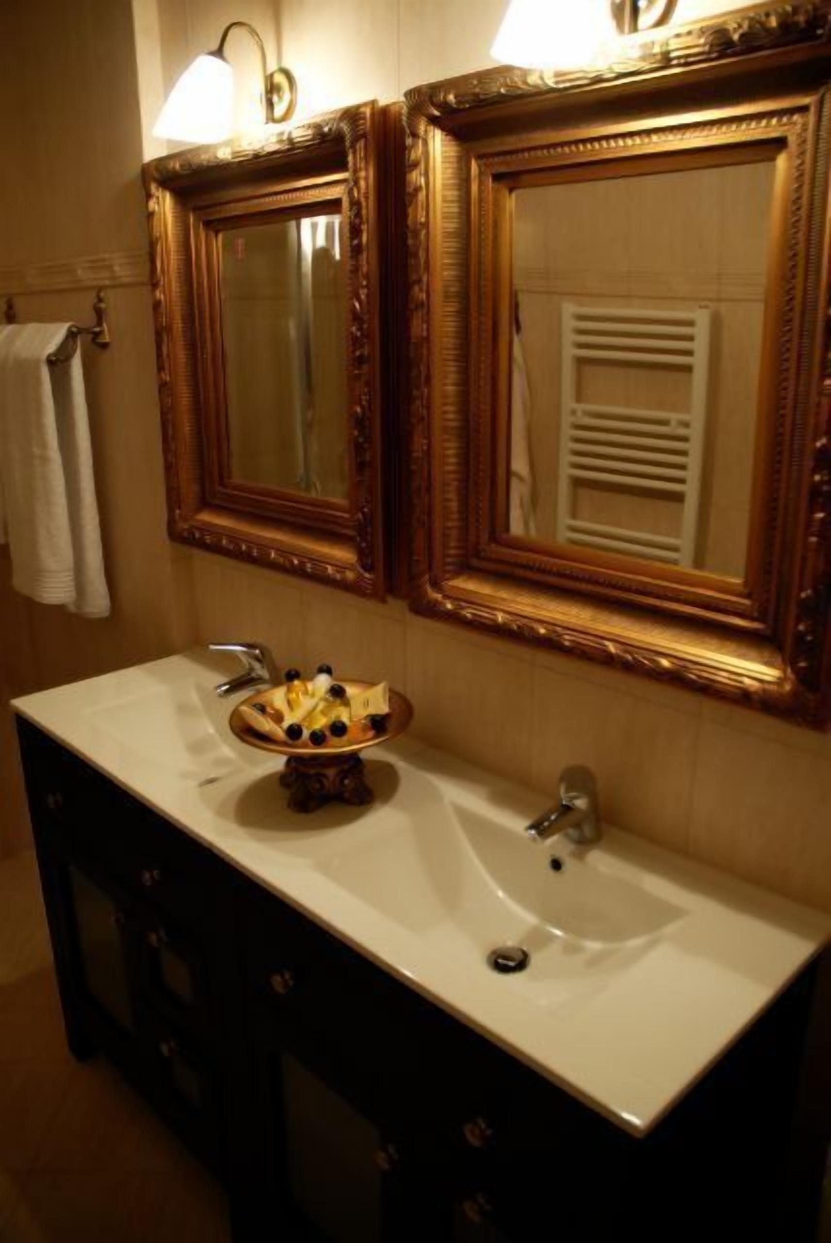 Une salle de bain avec deux miroirs richement décorés, un lavabo blanc et un bol décoratif posé sur le lavabo.