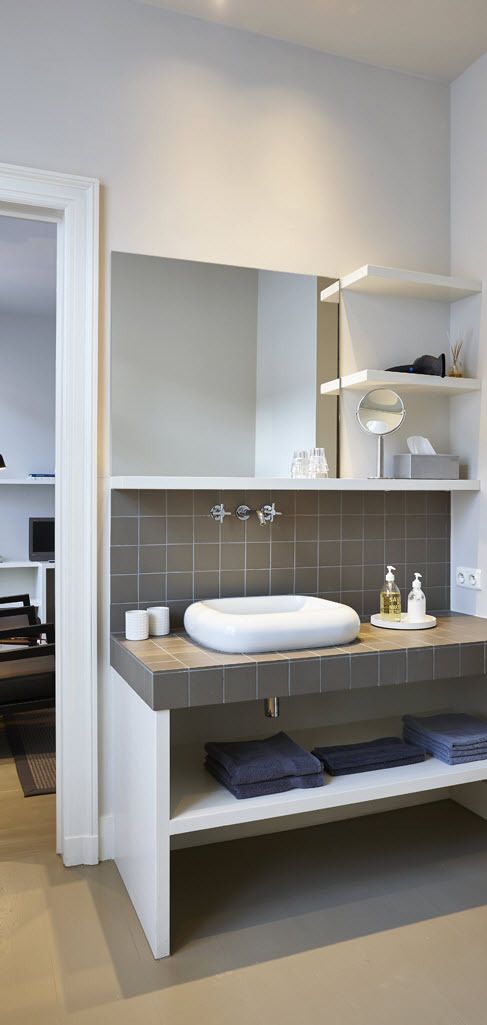 Une salle de bain moderne avec un lavabo blanc, des carreaux gris, un miroir et une étagère contenant des articles de toilette.