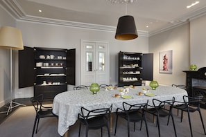 Uma sala de jantar com uma mesa redonda, cadeiras pretas e um lustre. Há prateleiras abertas com louças e um quadro na parede.