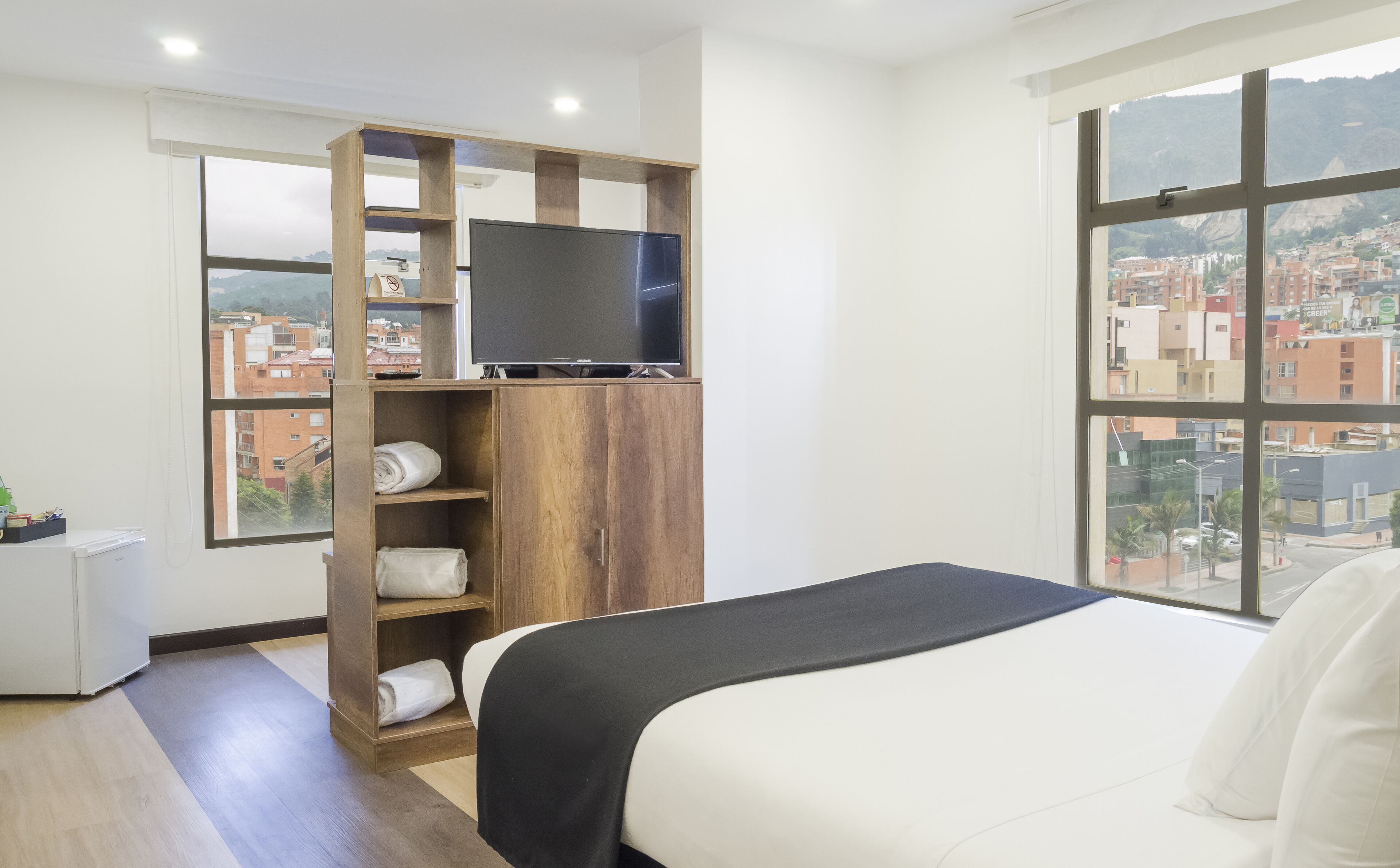 Habitación de hotel con cama, televisor en un estante de madera y vistas a la ciudad a través de grandes ventanales.