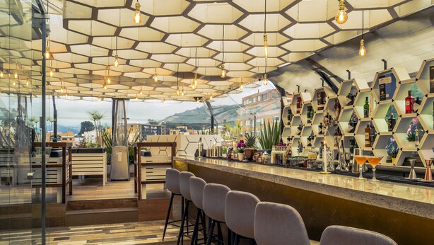 Zona de bar con paneles de techo hexagonales, taburetes y una exhibición de botellas y copas.