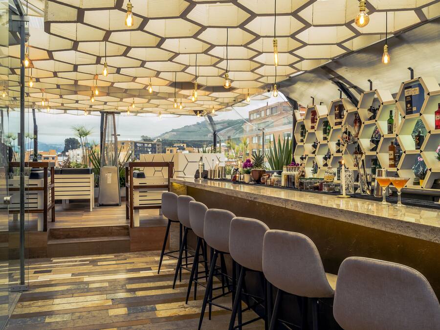 Uma área de bar com painéis hexagonais no teto, banquetas e uma seleção de garrafas e copos.