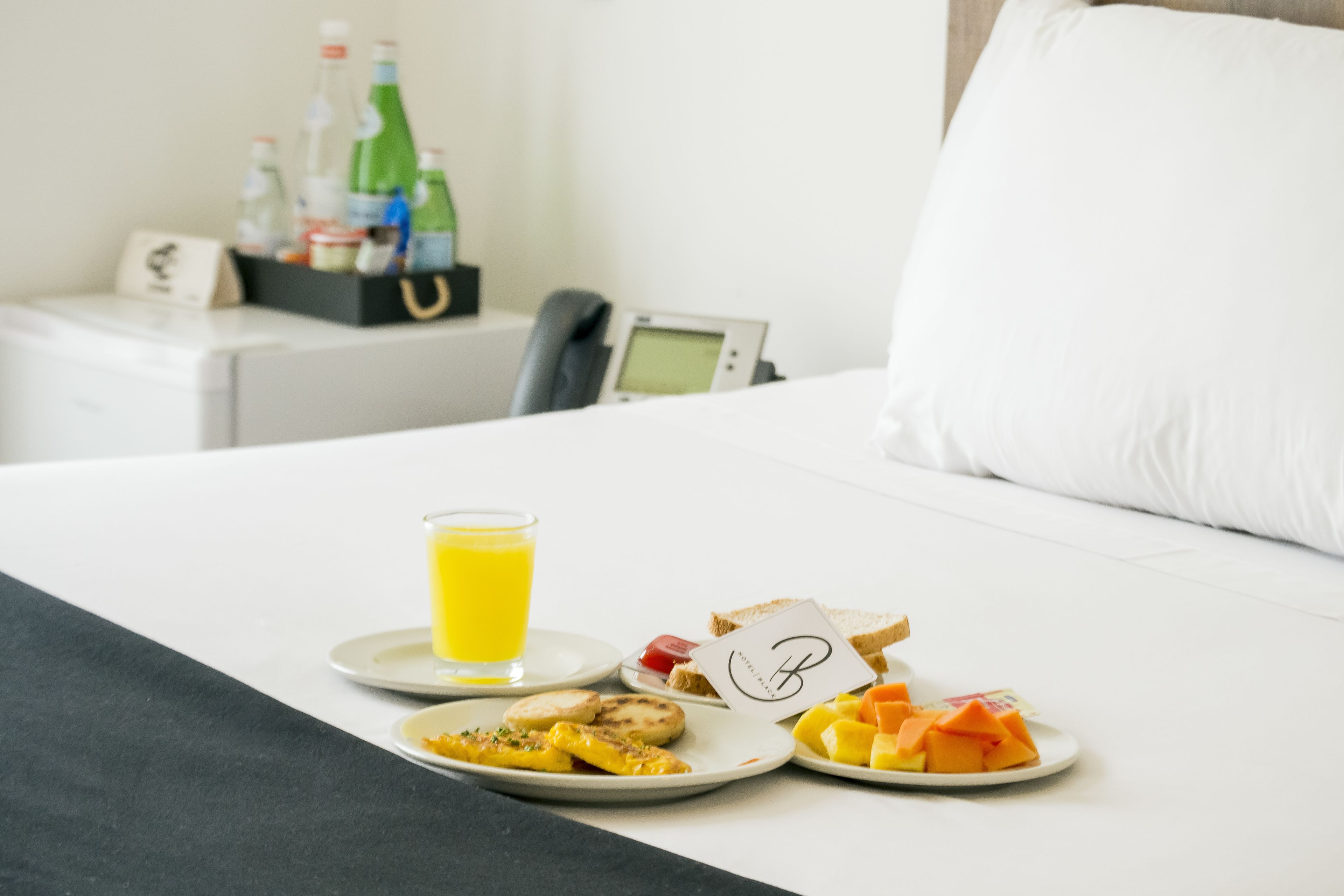 A neatly made bed with a pillow, a bedside table with a phone and a small screen, and a tray with breakfast items including toast, fruit, and a glass of orange juice.