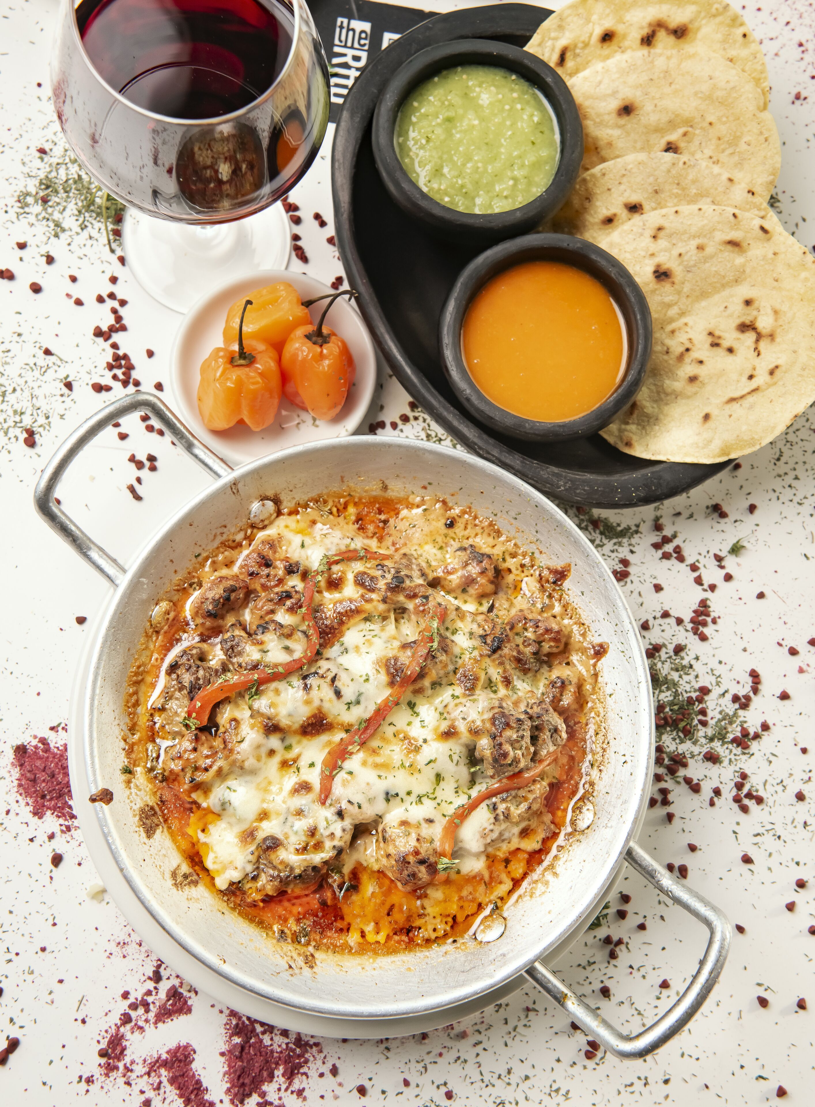 En måltid bestående av en gryta med mat, ett glas rött vin och en tallrik med tortillabröd, salsa och en grön sås.