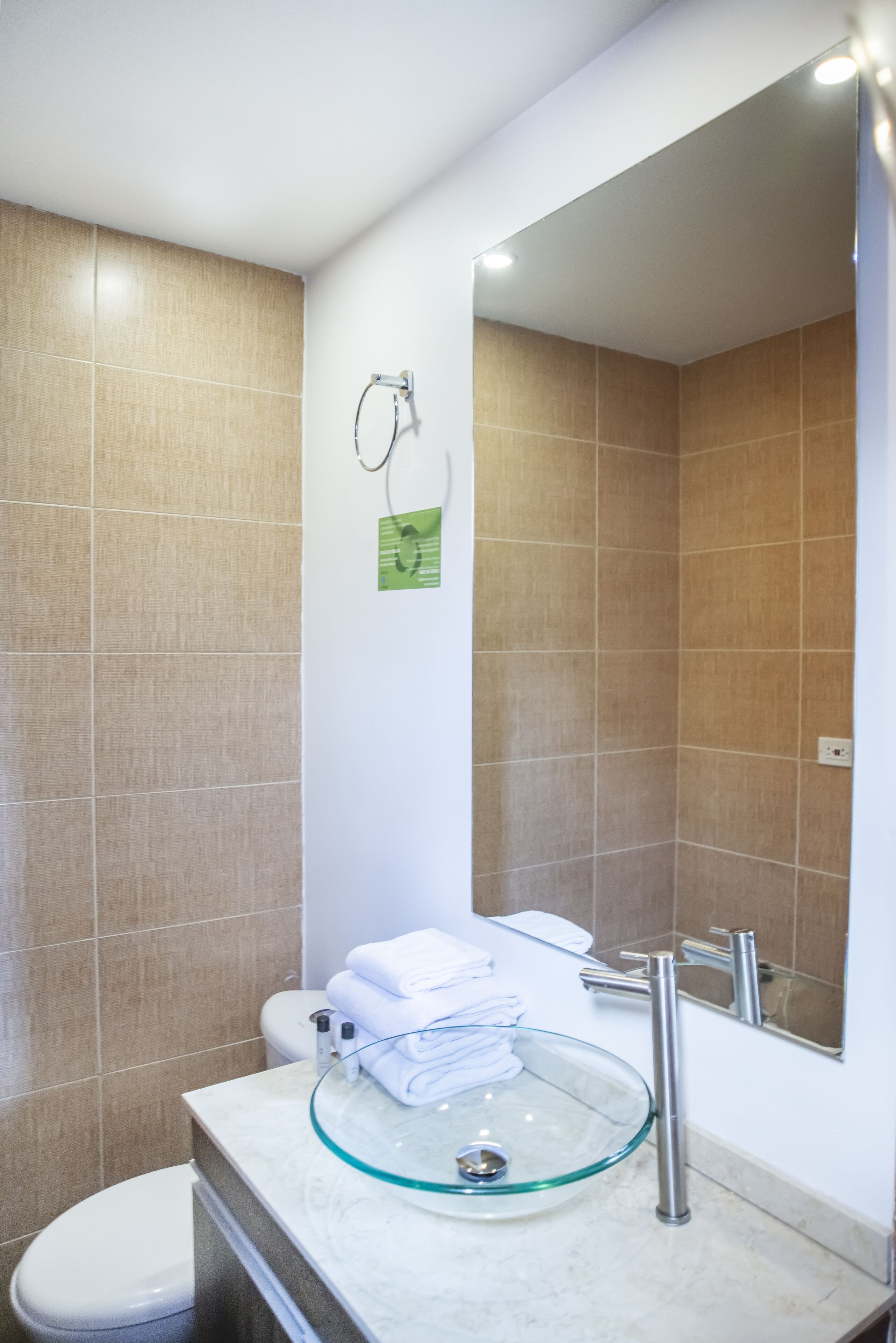 A bathroom with a glass sink, white toilet, and beige tiles.