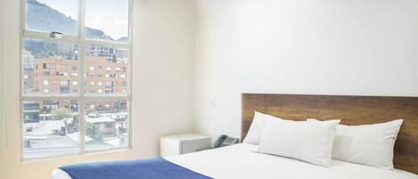 A hotel room with a large window, a bed with white bedding, a wooden headboard, and a bedside table with a phone.