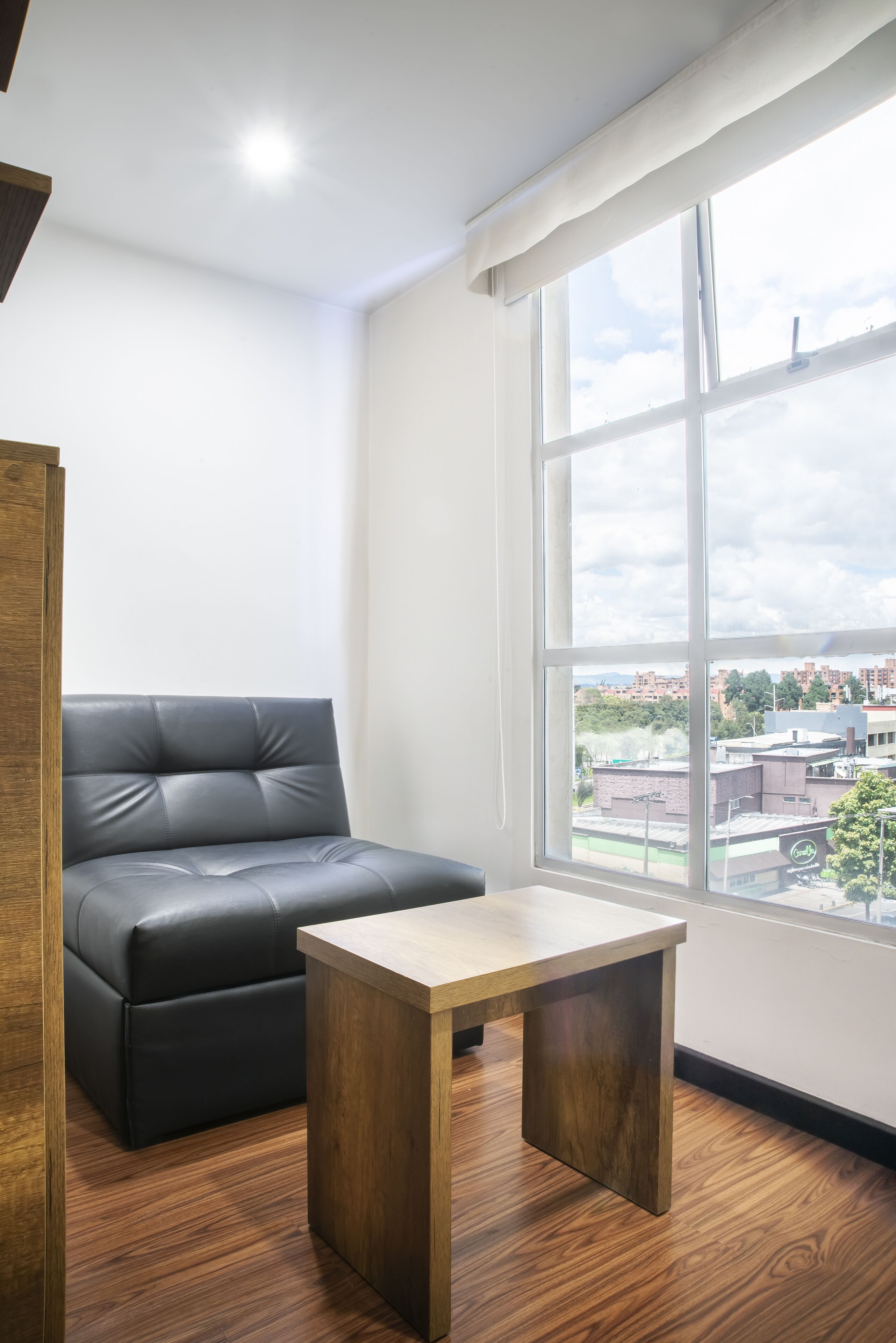 A modern room with a black leather sofa, a wooden coffee table, and a large window offering a city view.