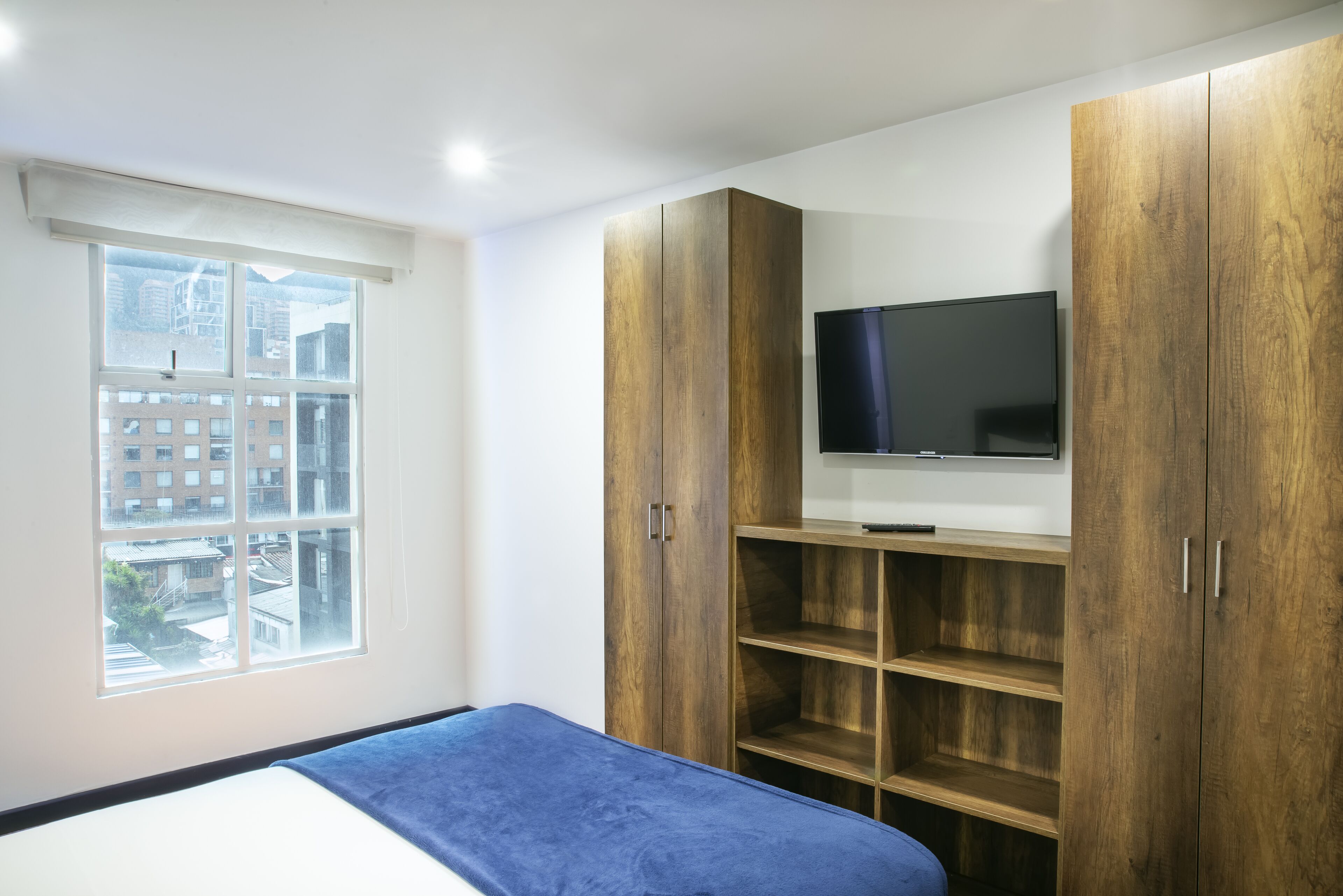 A bedroom with a bed, a TV mounted on wooden shelves, and a window with a view of buildings.