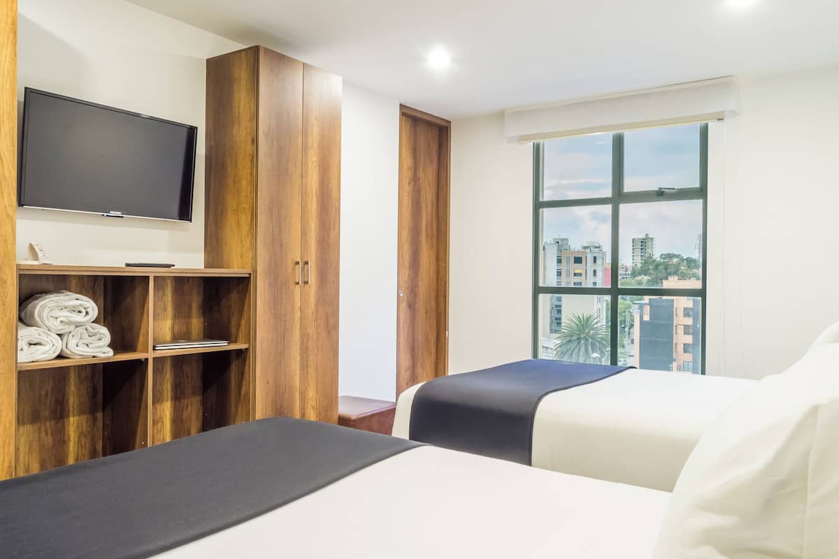 A hotel room with two beds, a TV on the wall, a large window with a city view, and a wooden wardrobe.