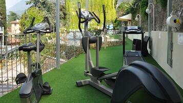 A gym with various exercise equipment, including treadmills and elliptical machines, on artificial grass under a covered area with a view of a mountainous landscape.
