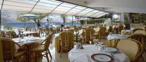 A restaurant with tables and chairs, a view of the sea, and a covered outdoor area.