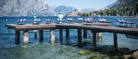 A pier with sun loungers extending into clear water, surrounded by rocky mountains and a distant town.