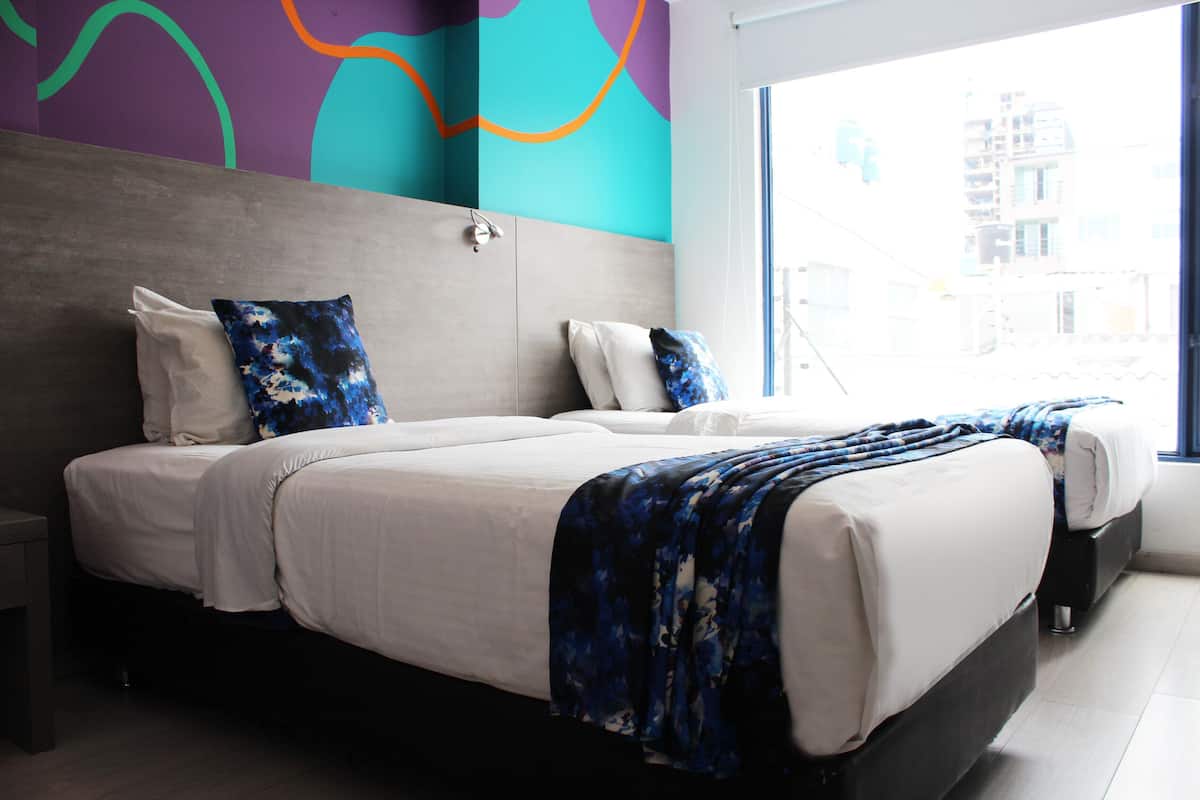 A modern hotel room with a large bed, a nightstand, and a view of the city through the window.