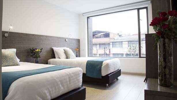 A modern hotel room with two beds, a large window, and a vase of red roses. - Agora Suites Self-Service Boutique Hotel (Bogotá)