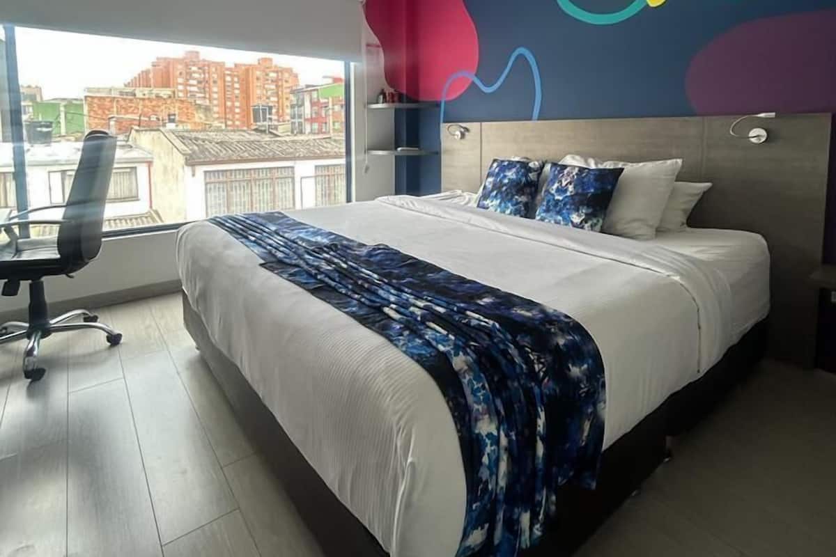 A modern bedroom with a large bed, a desk, and a view of the cityscape through large windows.