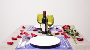 A table set with a bottle of wine, two wine glasses, a plate, a fork, a knife, and scattered rose petals. - Agora Suites Self-Service Boutique Hotel (Bogotá)