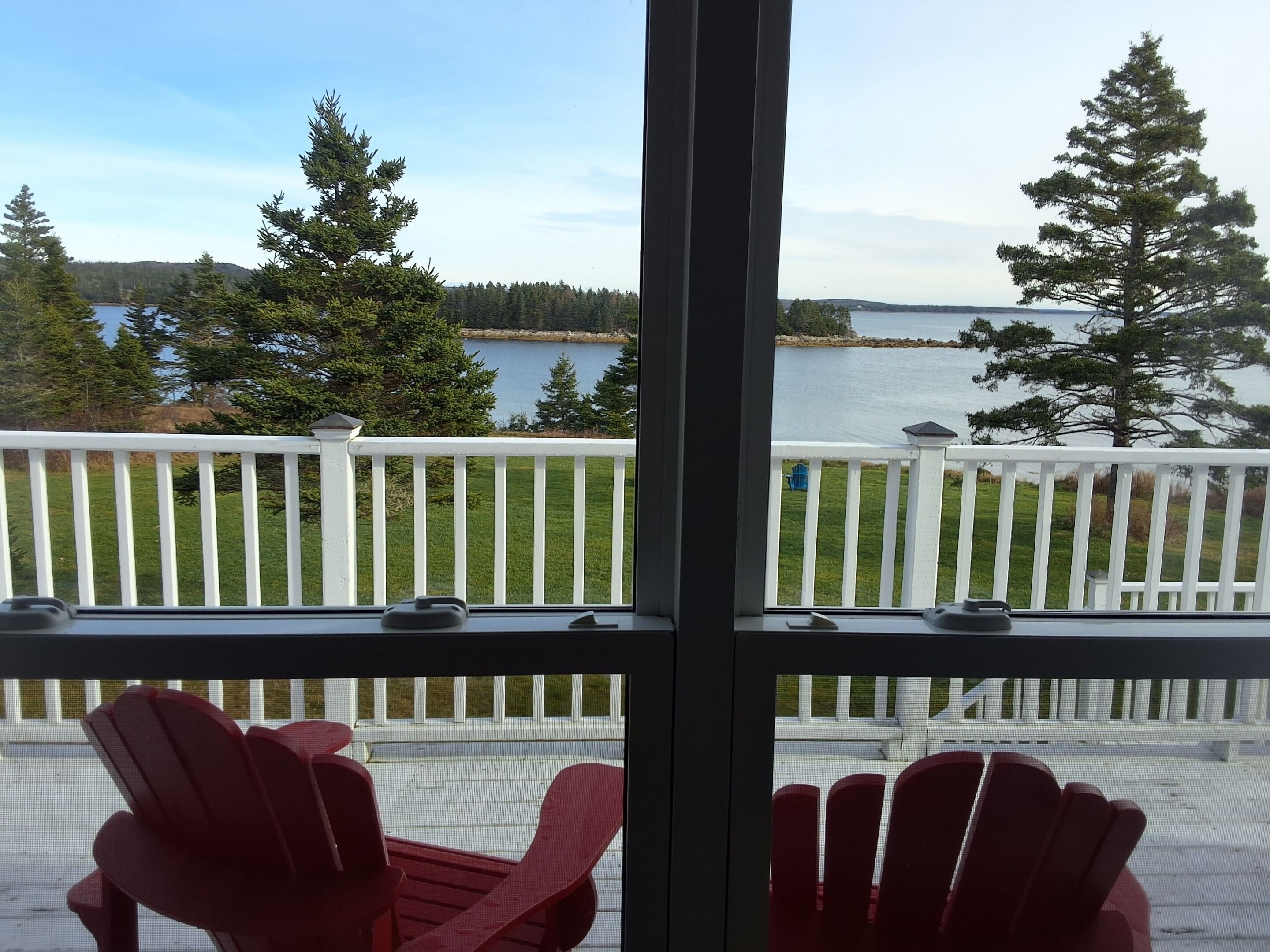 A balcony with two red Adirondack chairs, a white railing, and a view of a lake and forest.