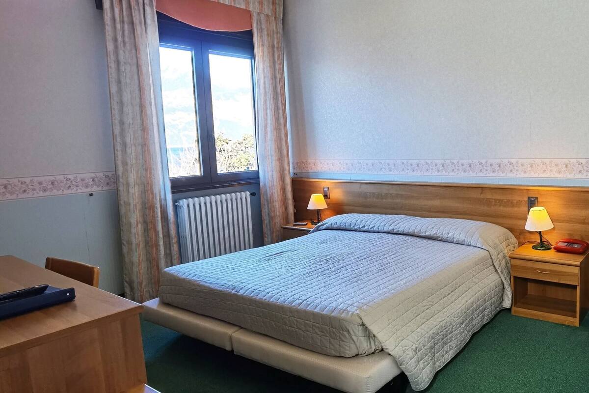 A hotel room with a bed, bedside tables, a window with curtains, a radiator, and a desk with a chair.