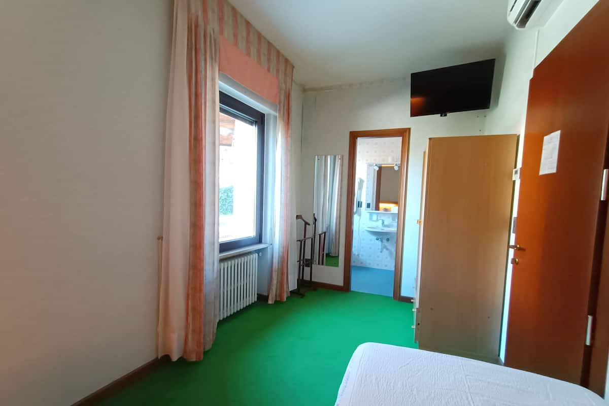 A hotel room with a green carpet, a bed, a window with curtains, a radiator, a TV mounted on the wall, and a door leading to a bathroom.