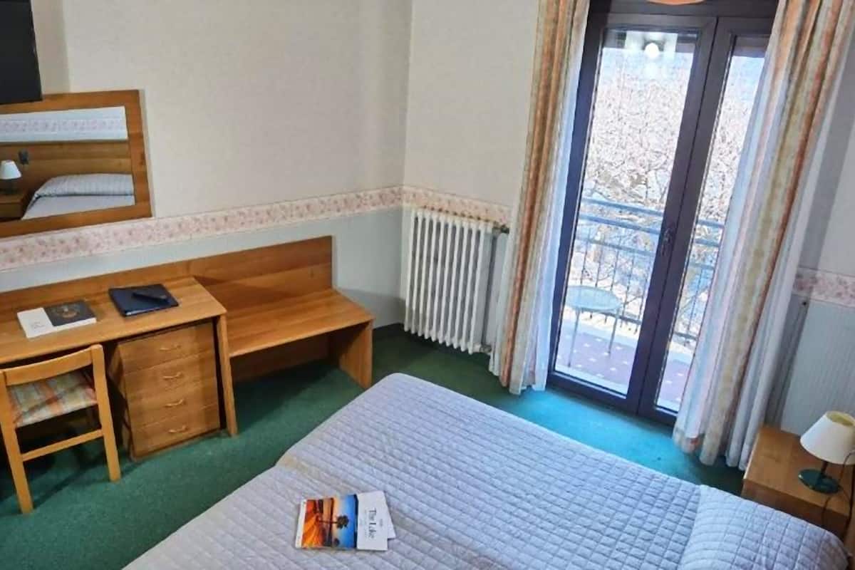 A hotel room with a bed, a wooden desk, a chair, a radiator, and a window with curtains.