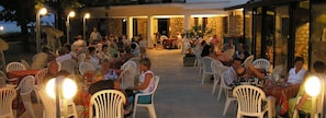 Un espace extérieur aménagé pour les repas, avec des chaises et des tables en plastique blanc, des murs en pierre et un bâtiment en pierre en arrière-plan.