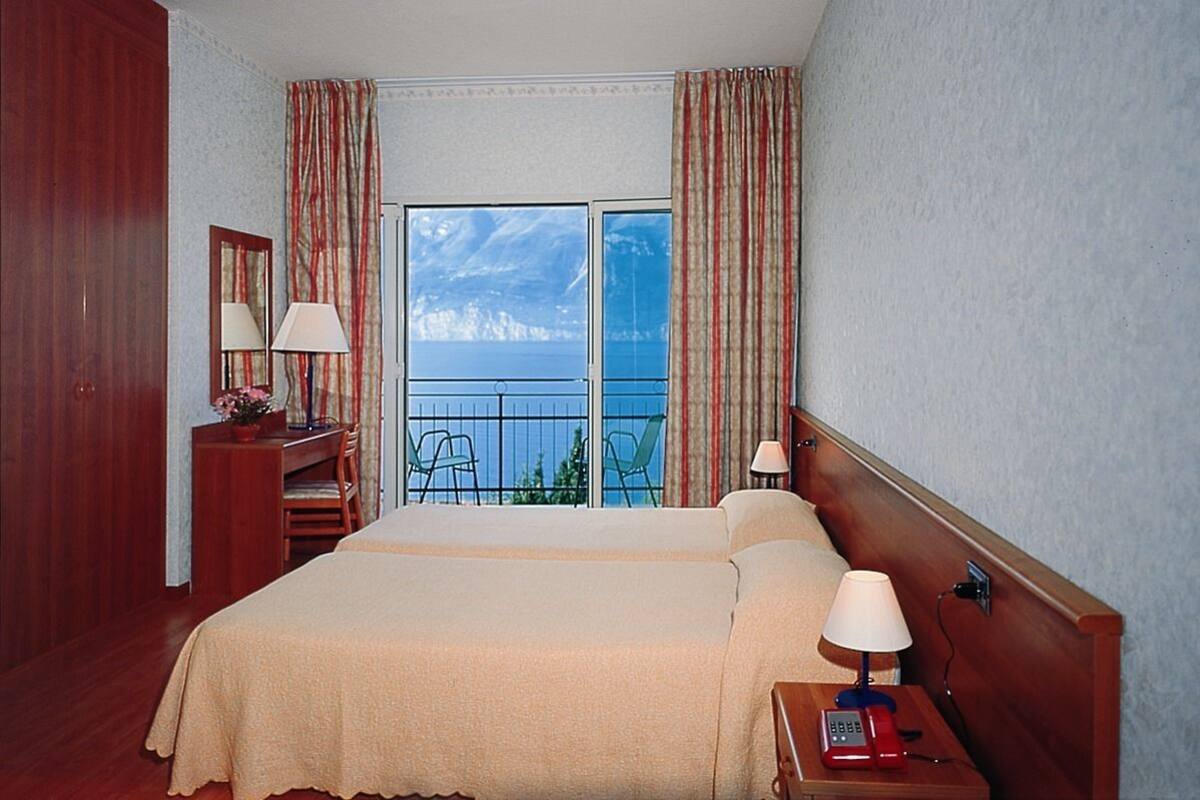 Een hotelkamer met een groot bed, een bureau met stoel, een telefoon en uitzicht op de zee vanaf een balkon.