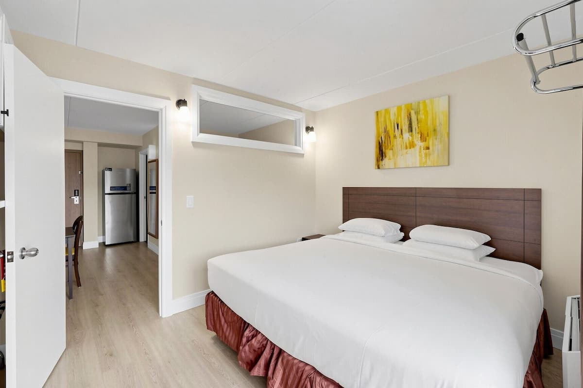 Habitación de hotel con una cama, una puerta que da a un pasillo, un refrigerador y una lámpara fijada a la pared.