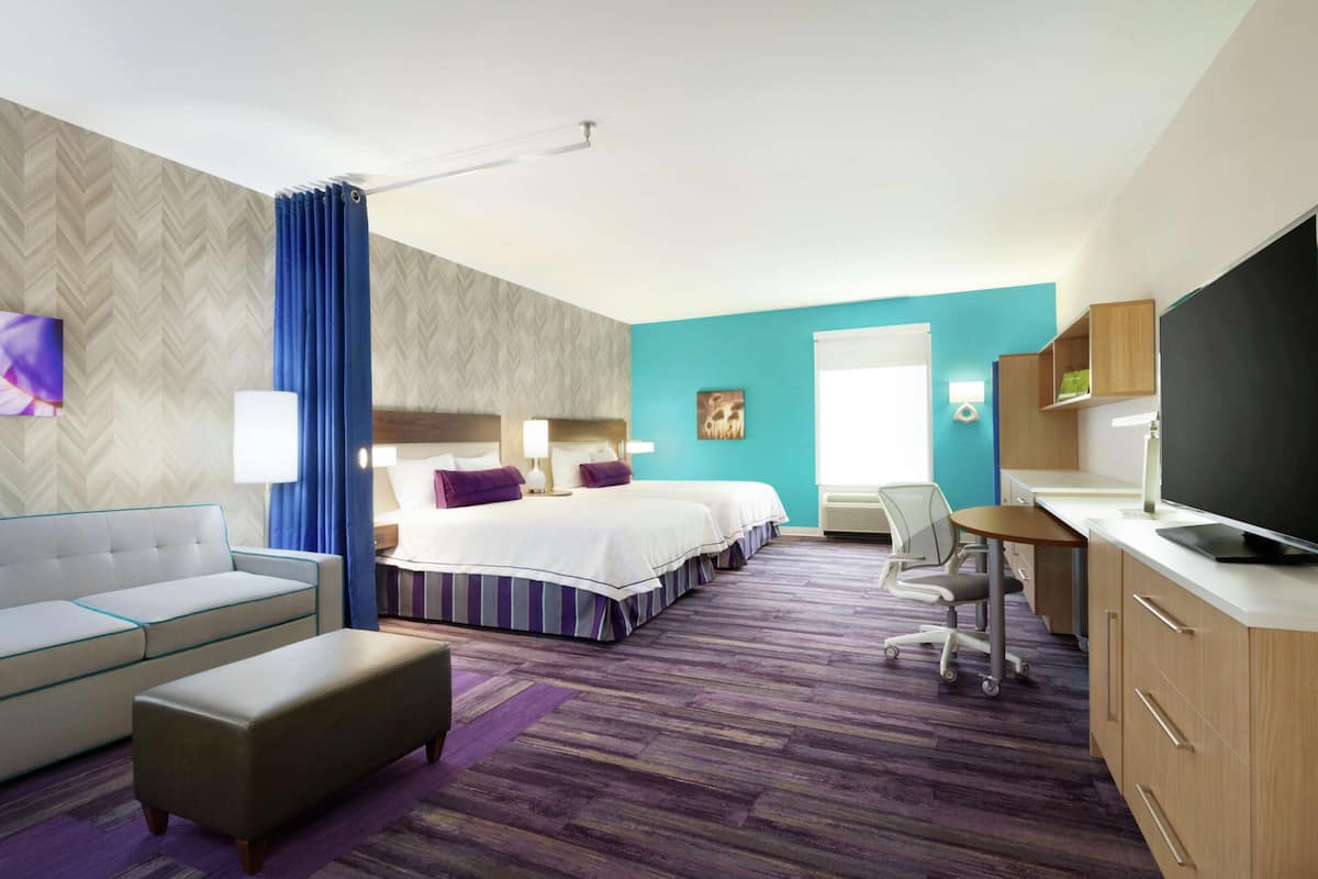 A hotel room with a sofa, two beds, a desk, a TV, and a bathroom.