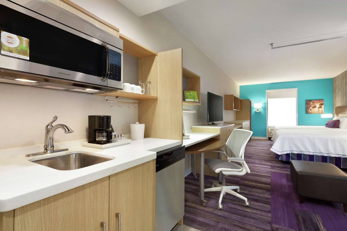 A modern kitchen with a microwave, coffee maker, and dishwasher. There is a dining area with a chair and a bed in the background.
