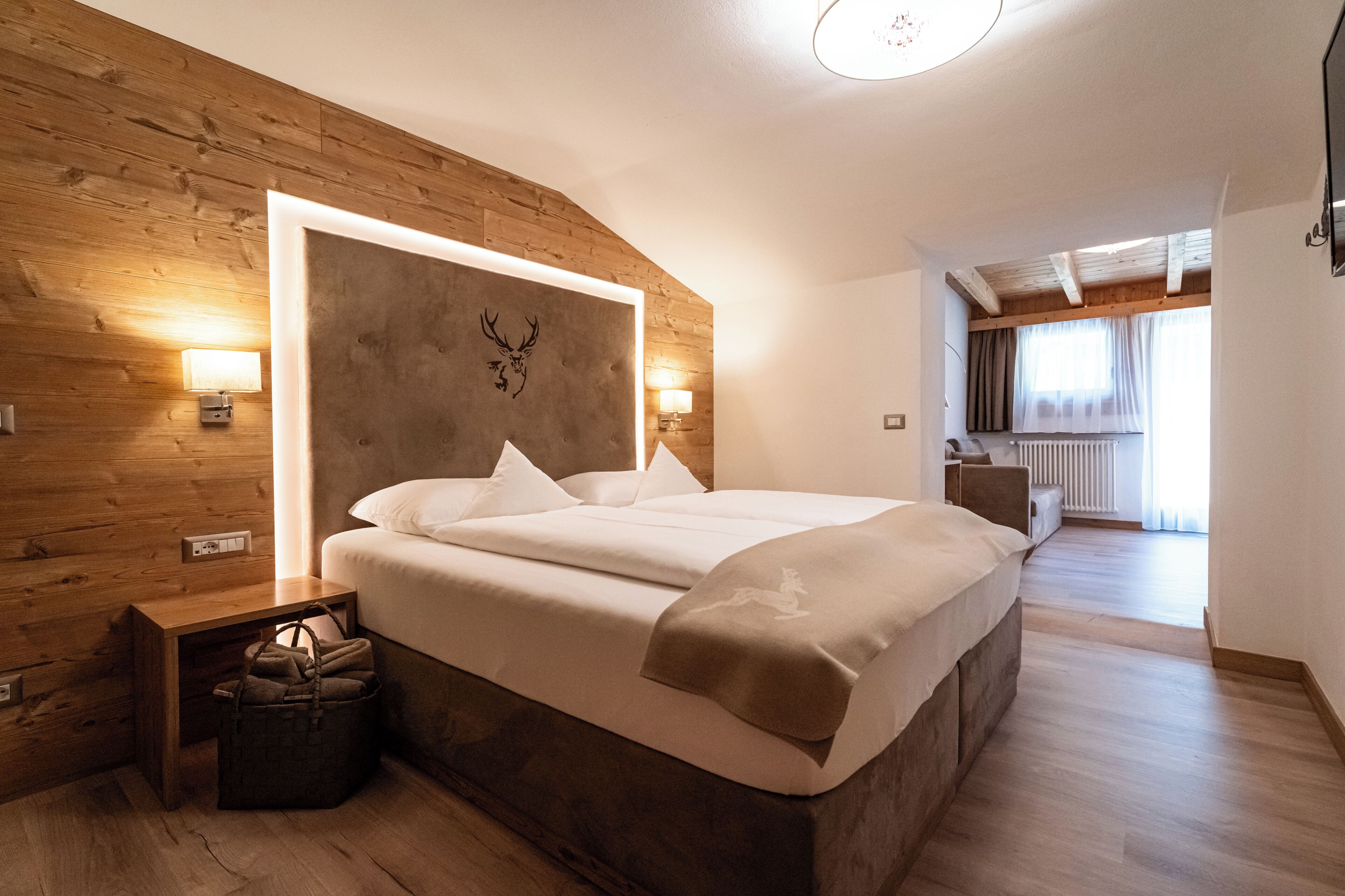 Une chambre à coucher avec une tête de lit en bois, un lit recouvert de draps blancs, des lampes de chevet et un sac posé sur le sol.