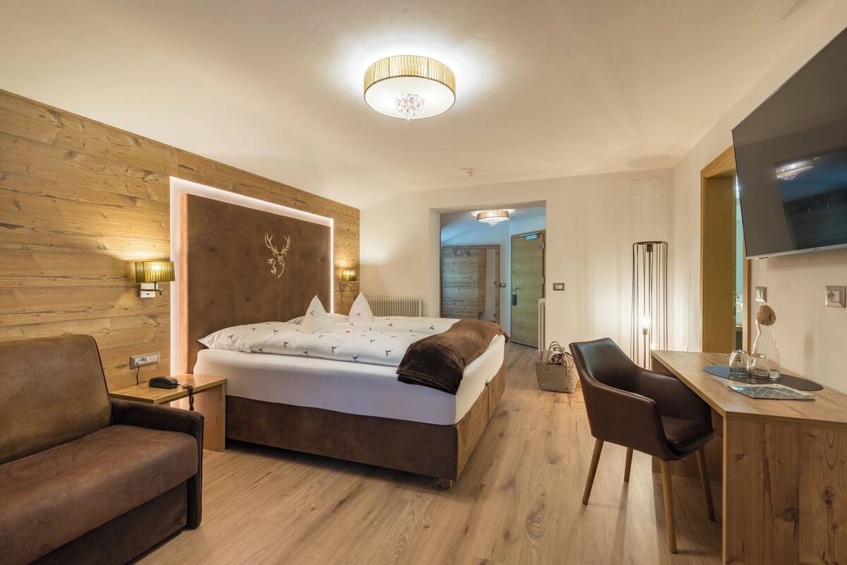 A modern hotel room with a wooden headboard, a bed with white linens, a desk with a chair, and a large mirror.