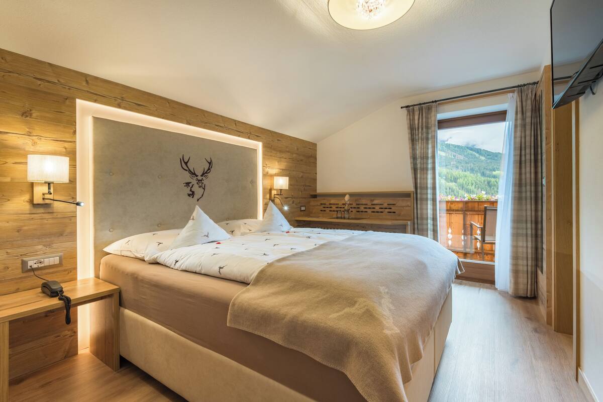 A bedroom with a large bed, wooden headboard, bedside lamps, and a view of a balcony with outdoor scenery.