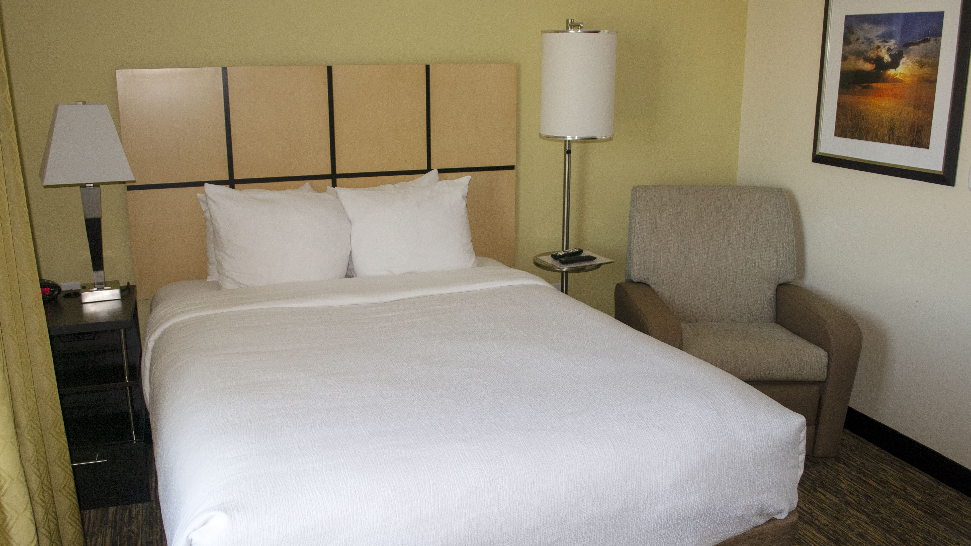 A hotel room with a large bed, a chair, a nightstand with a lamp, and a framed picture on the wall.