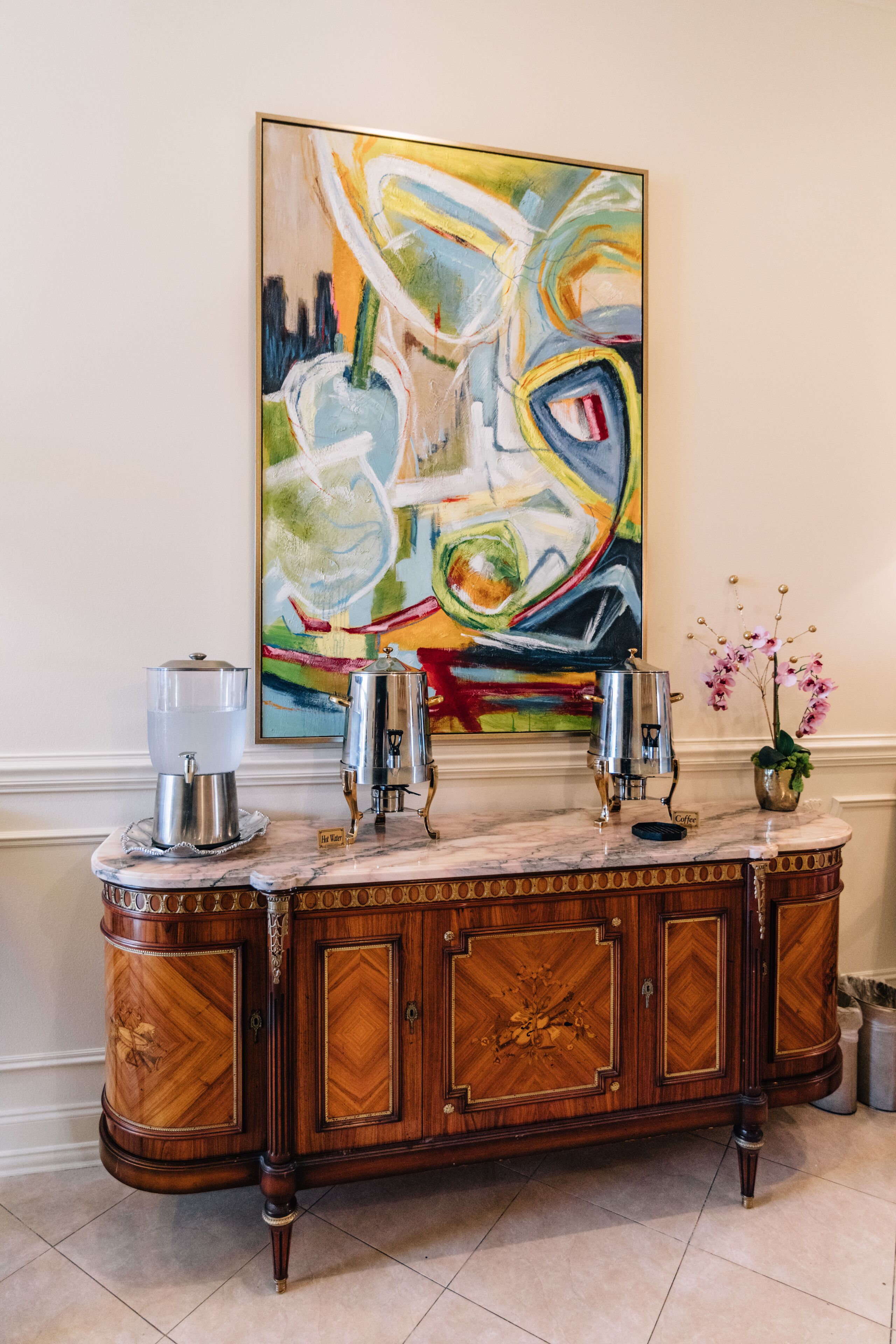 A marble countertop with two coffee makers, a decorative vase with orchids, and a colorful abstract painting on the wall.