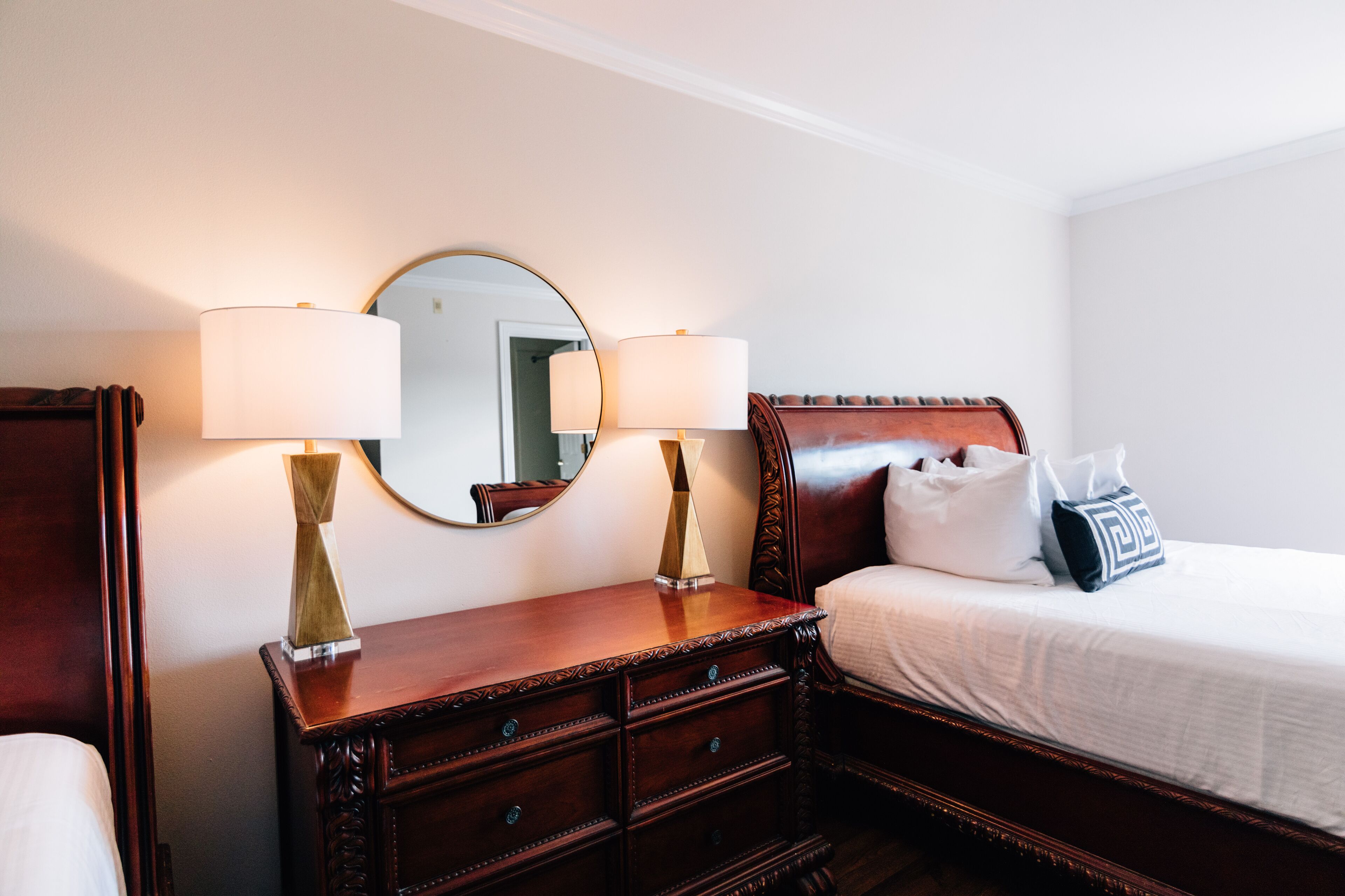 A hotel room with a wooden dresser, two bedside lamps, a mirror, a bed with white bedding, and a pillow with a geometric pattern.