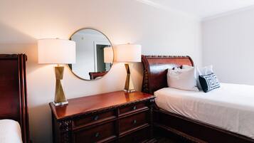 A hotel room with a wooden dresser, two bedside lamps, a mirror, a bed with white bedding, and a pillow with a geometric pattern.