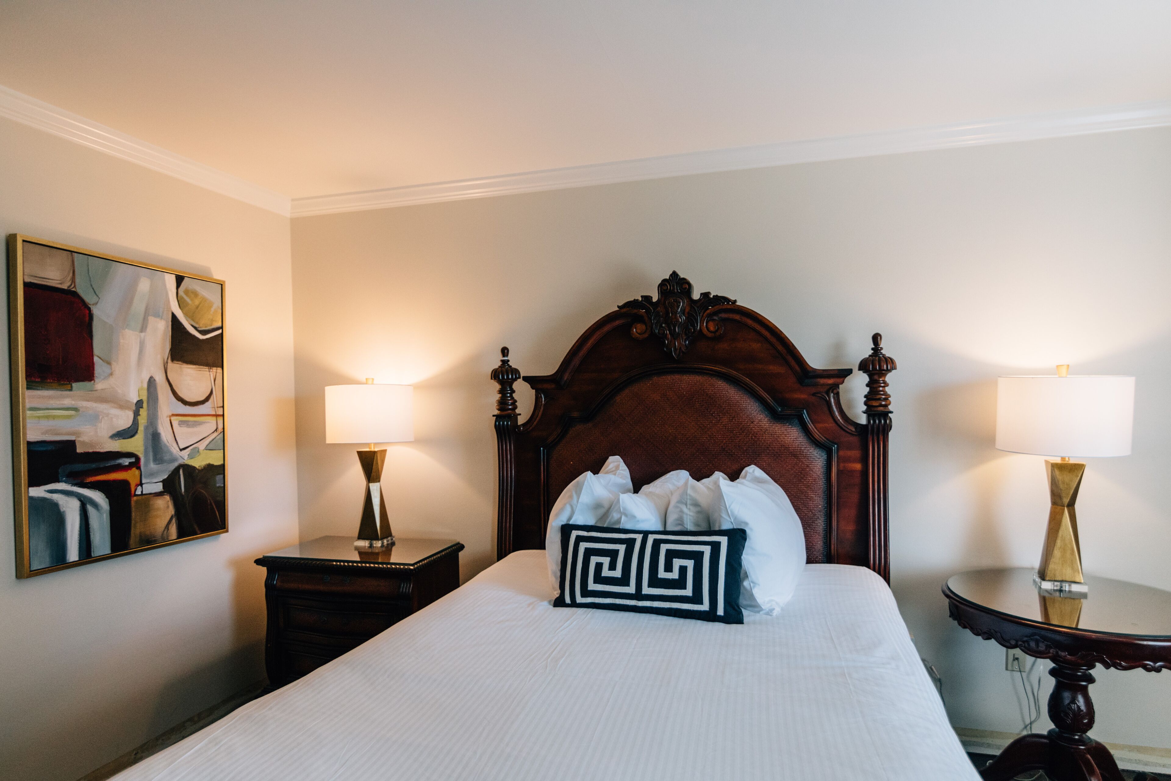 A neatly made bed with a wooden headboard, two bedside tables with lamps, and a framed abstract painting on the wall.