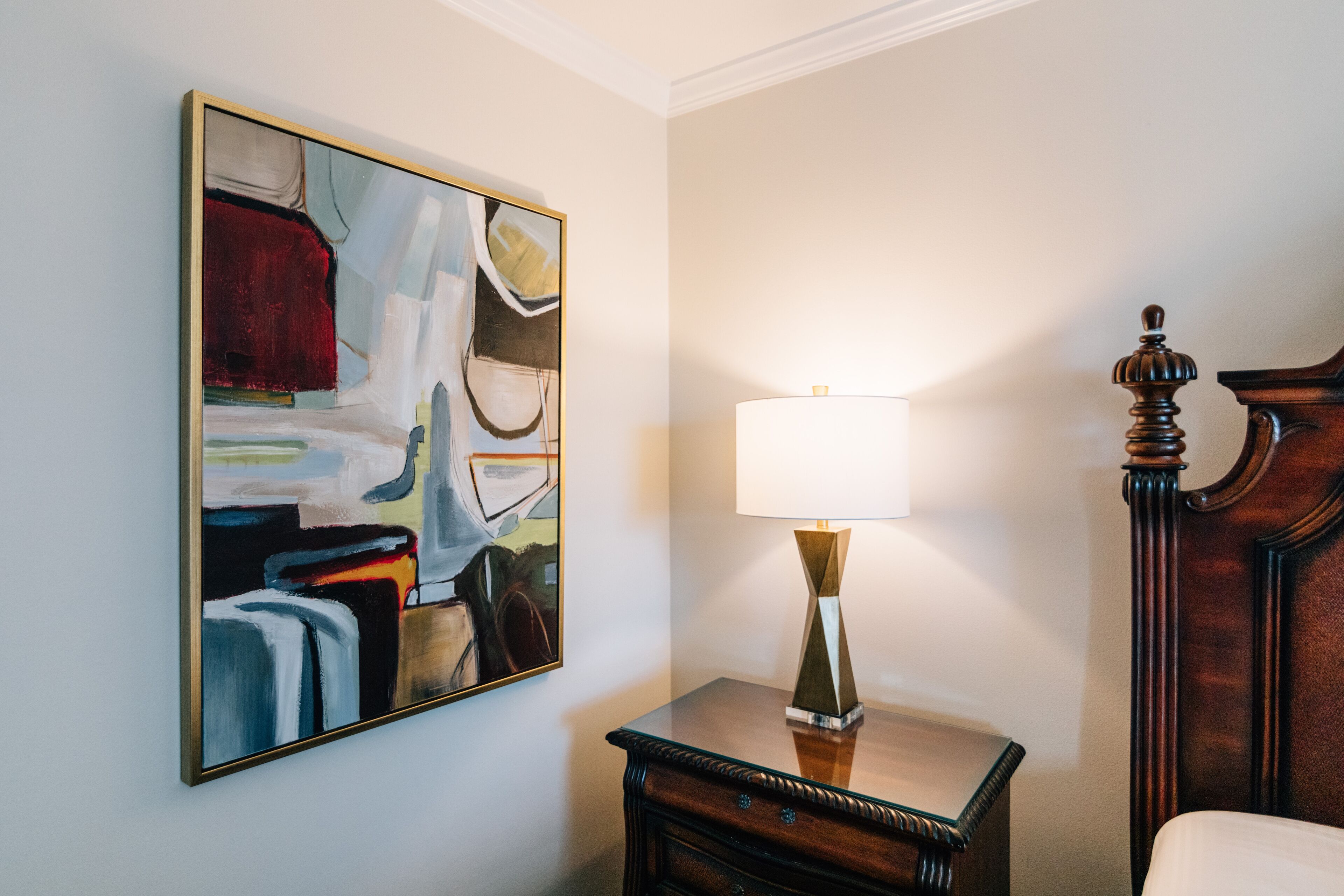 A room with a painting on the wall, a bedside table with a lamp, and a wooden headboard.