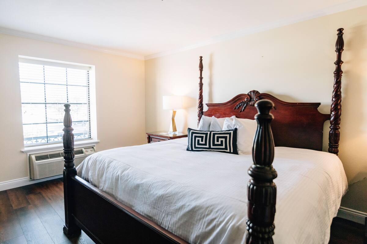 A neatly made bed with a wooden headboard, a bedside table with a lamp, and a window with blinds.