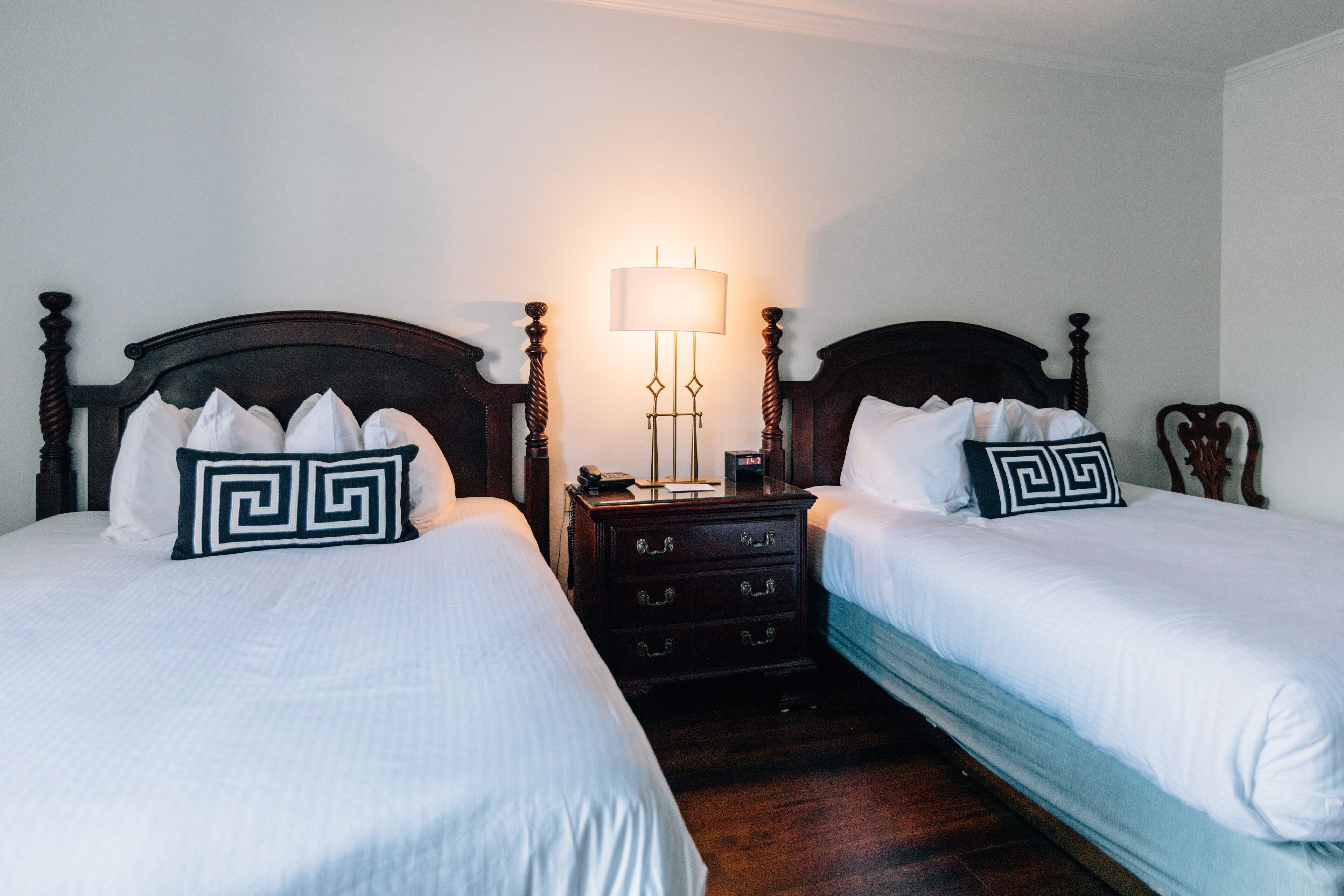 Quarto de hotel com duas camas, cada uma com roupa de cama branca e almofadas geométricas pretas. Há uma mesa de cabeceira com uma lâmpada e um telefone.