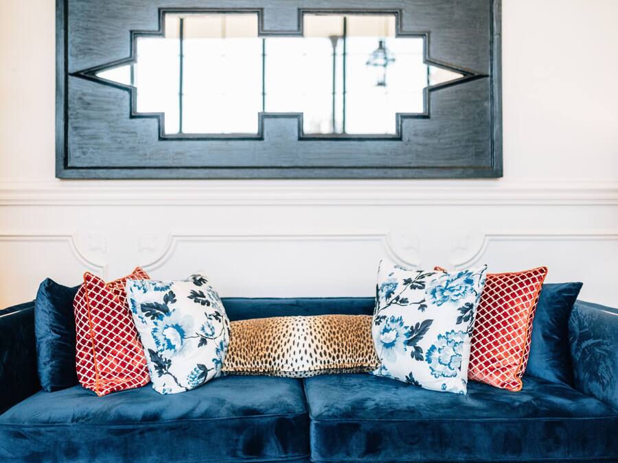 A dark blue velvet sofa with floral and patterned cushions, a geometric wall art piece above, and a white wall with a decorative molding.