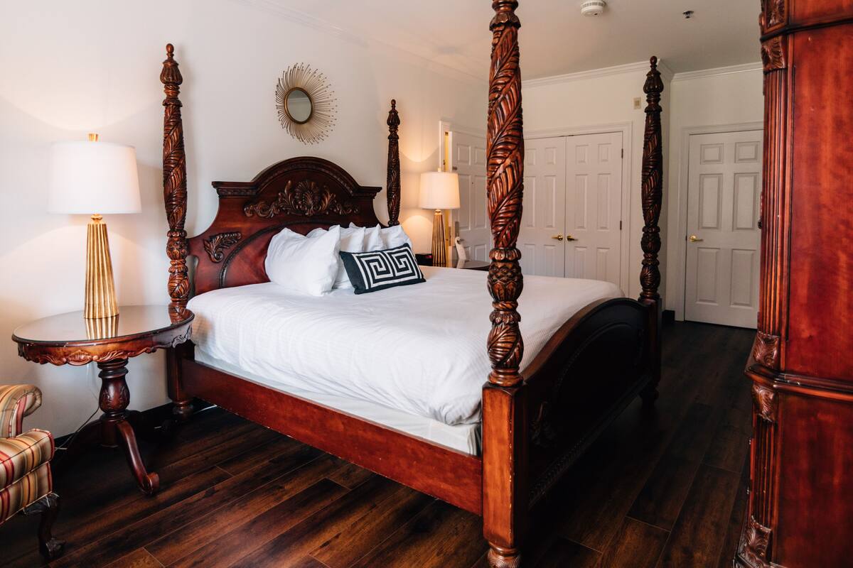 A four-poster bed with a wooden headboard, two bedside tables with lamps, a chair, and a mirror on the wall.