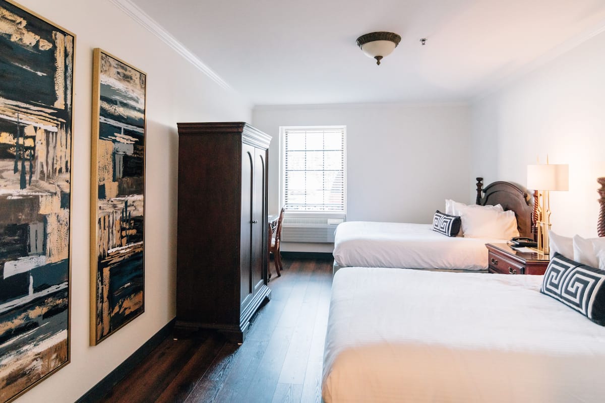 A hotel room with two beds, a large window, a wardrobe, and artwork on the walls.