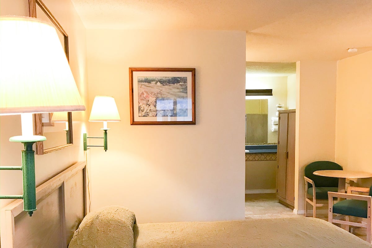 A hotel room with a bed, a desk, a chair, a lamp, a framed picture on the wall, and a bathroom visible through an open door.