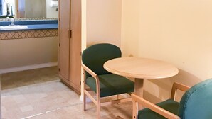 Una pequeña kitchenette con un lavamanos, un espejo y un armario. Hay dos sillas verdes y una mesita redonda.
